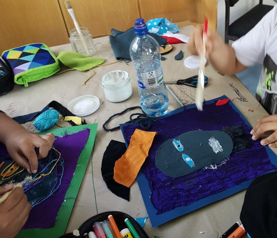 Bild: Auf einem Tisch liegen Kleber und Stoffe mit denen Kinder an einer Collage arbeiten. Die Collage hat die Form eines Gesichts.
