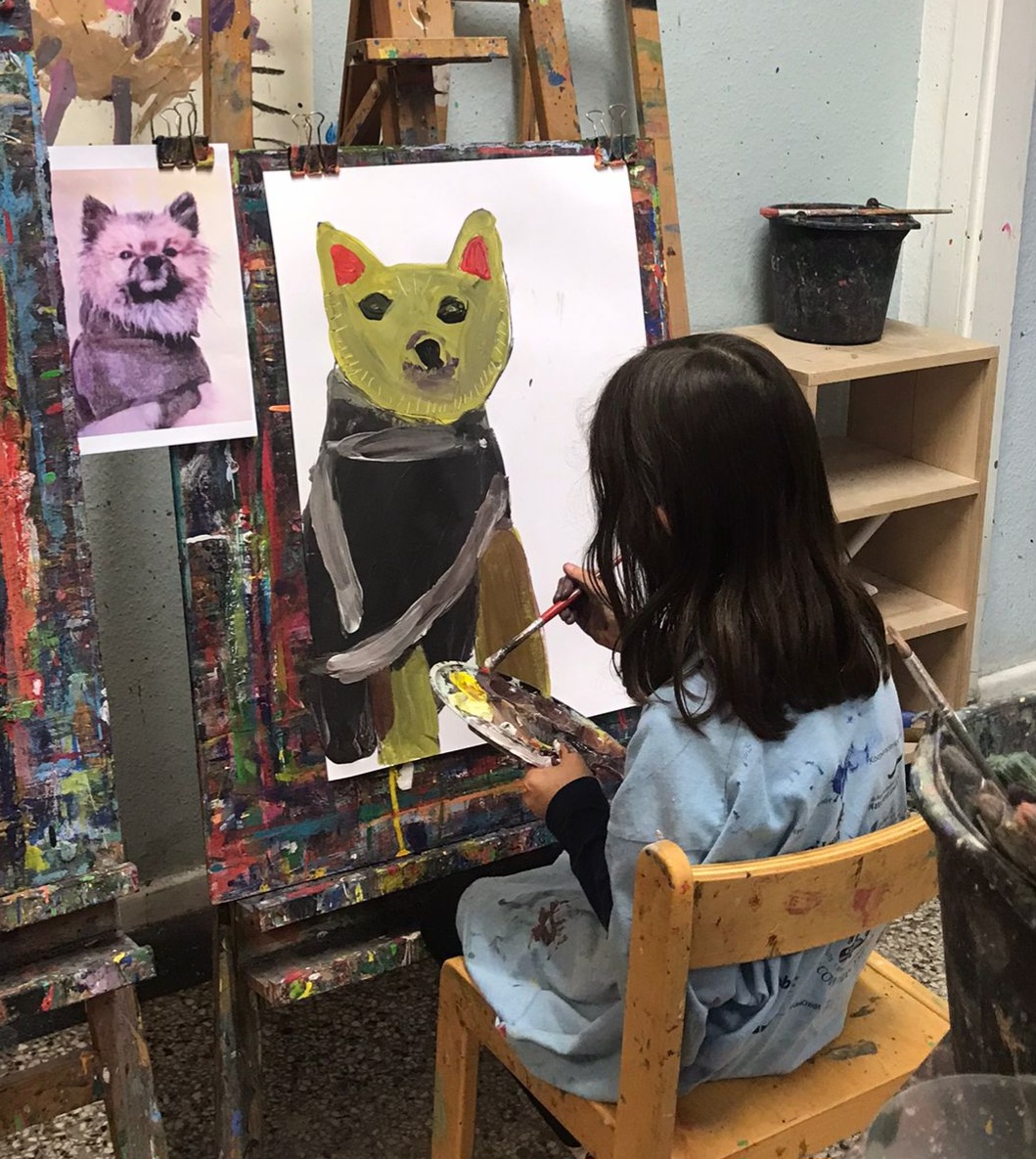 Das Foto zeigt ein junges Mädchen mit braunen Haaren, das vor einer Staffelei sitzt und einen Hund malt, anhand eines daneben hängenden Fotos