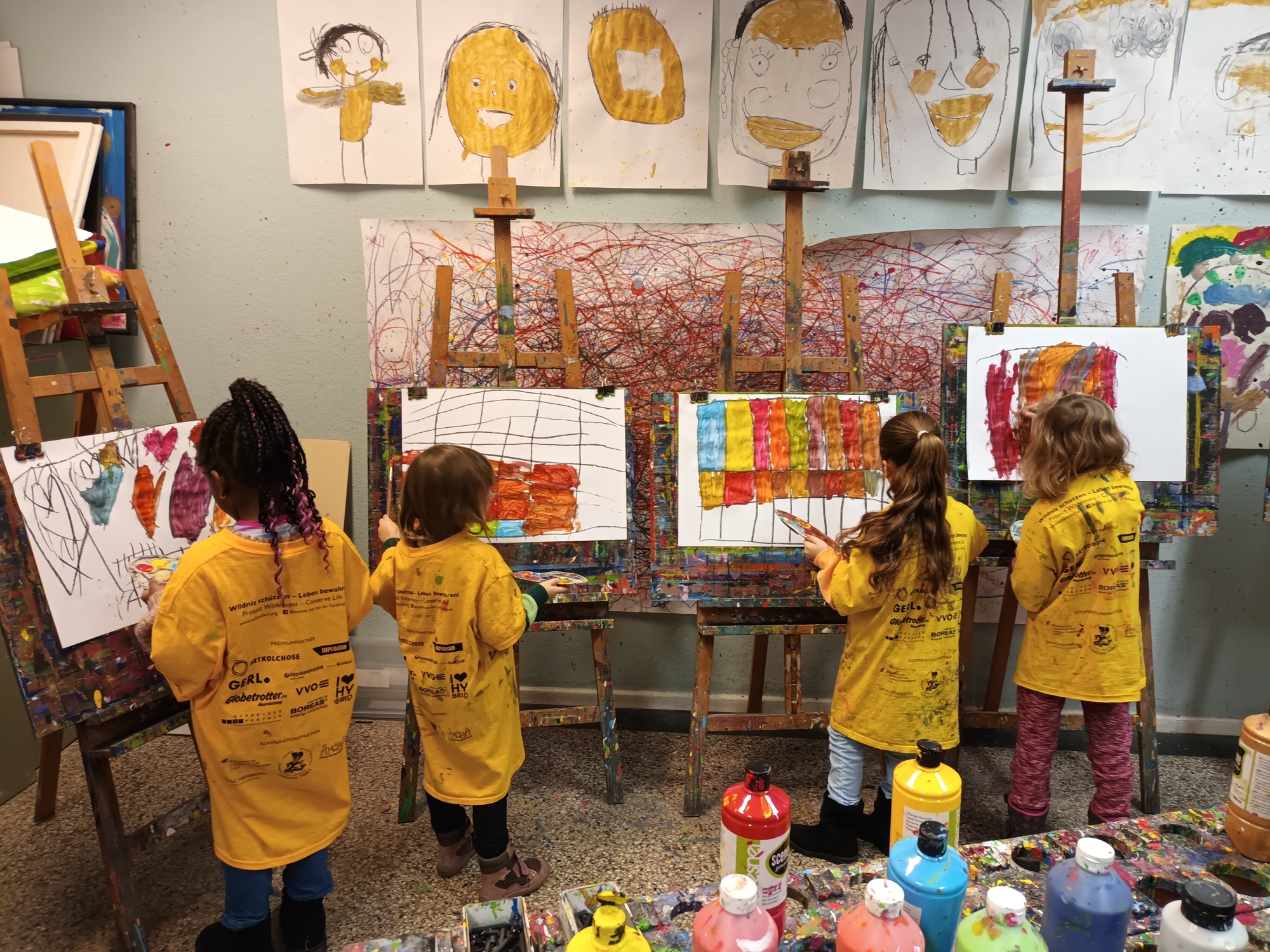 Beispielfoto, zu sehen sind 4 Mädchen, alle 4 stehen vor Staffeleien und Malen mit bunten Farben verschiedene Bilder