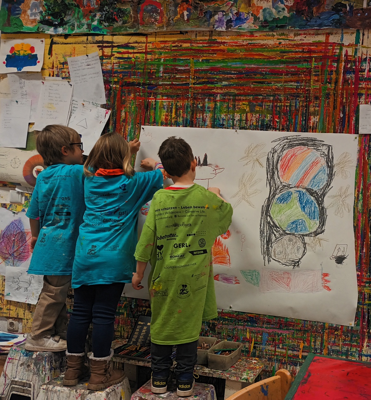 Beispielfoto, zu sehen sind drei Kinder, die vor einem großen Blatt Papier stehen, welches an der Wand hängt und gemeinsam Figuren malen