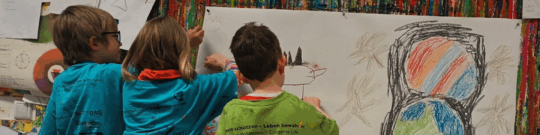 Drei Kinder stehen vor einer großen Leinwand und malen auf diese.